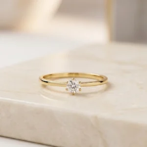 Anillo solitario dorado para compromiso sobre base beige de joyería premium, disponible en Luxor SM Joyería Santa Marta.