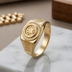 Anillo masculino dorado con corona grabada sobre base clara de joyería premium, disponible en Luxor SM Joyería Santa Marta.