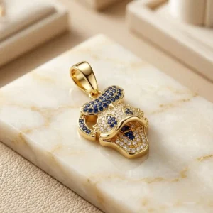 Dije dorado tipo personaje con detalles azules y brillantes sobre base de mármol claro, joya exclusiva de Luxor SM Joyería en Santa Marta