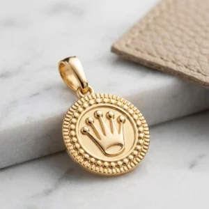 Dije dorado redondo con corona en relieve sobre base de mármol claro, joya elegante de Luxor SM Joyería en Santa Marta.