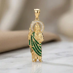 Dije de San Judas Tadeo dorado con detalles verdes y halo brillante sobre base de mármol claro, joya religiosa de Luxor SM Joyería en Santa Marta.