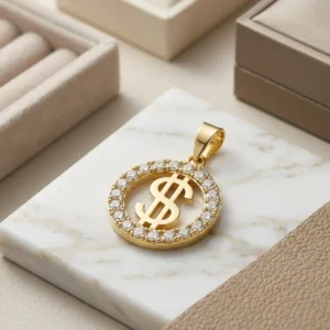 Dije circular dorado con signo de dólar y borde brillante sobre base de mármol claro, joya elegante de Luxor SM Joyería en Santa Marta.