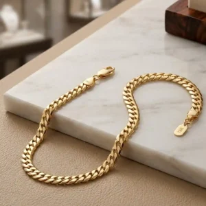 Pulsera cubana plana dorada sobre base de mármol claro, joya elegante de Luxor SM Joyería en Santa Marta.
